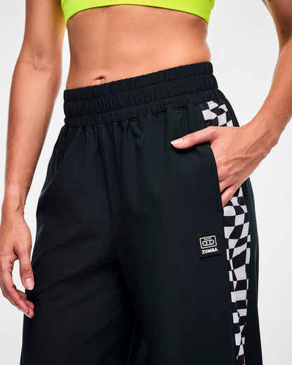 Close-up of the "ZUMBA" cassette black and white logo and side pocket of the black track pants with checkerboard side panel.