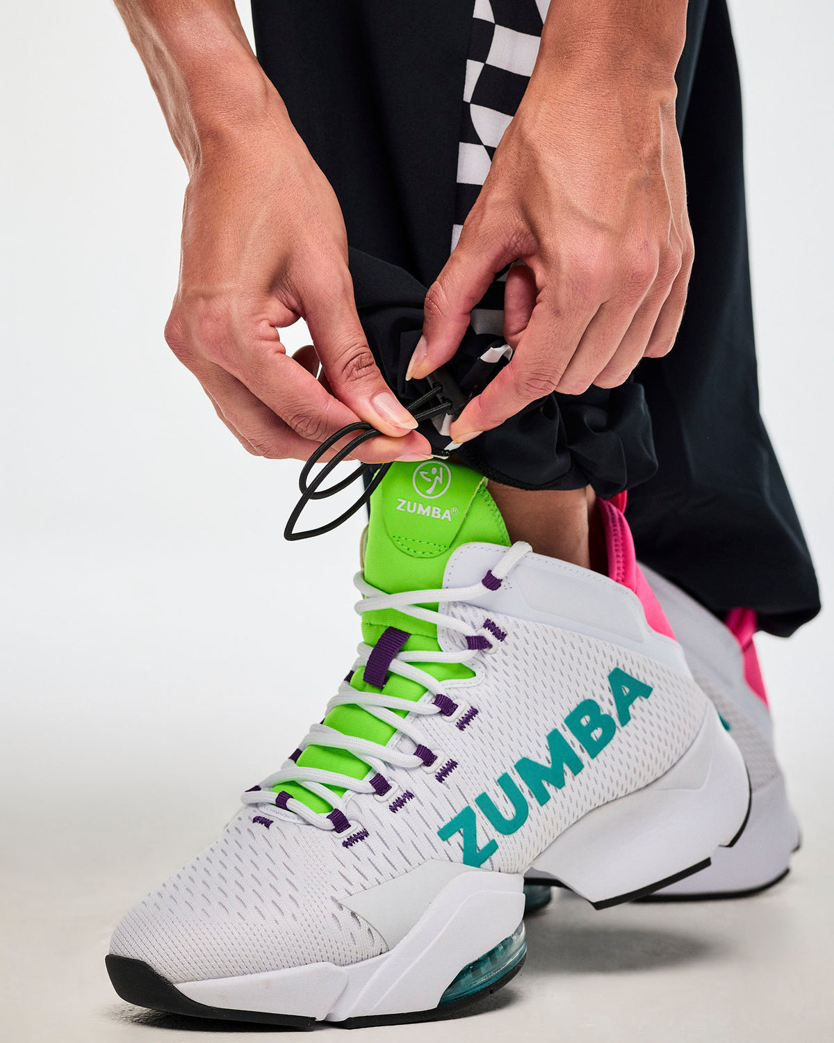 Close-up of a female model adjusting the bungee cords at the bottom leg opening and showing the white "ZUMBA" sneakers with green and purple highlights.
