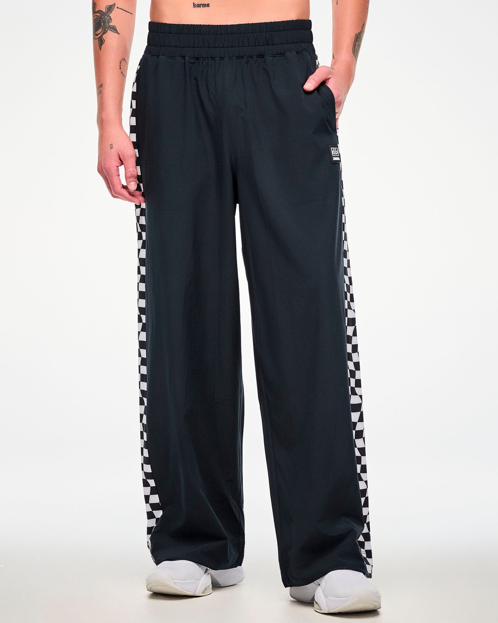 Male model wearing black track pants with a checkerboard side panel and white sneakers, hand in the side pocket.