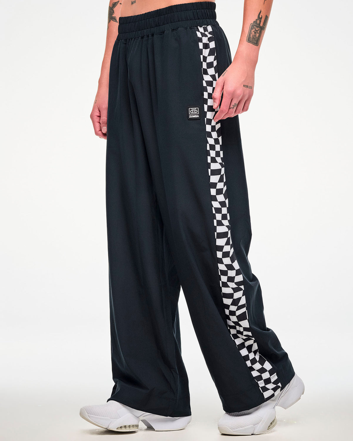 Shown from the back, male model wearing black wide-leg track pant with a checkerboard side panel and paired with white sneakers.