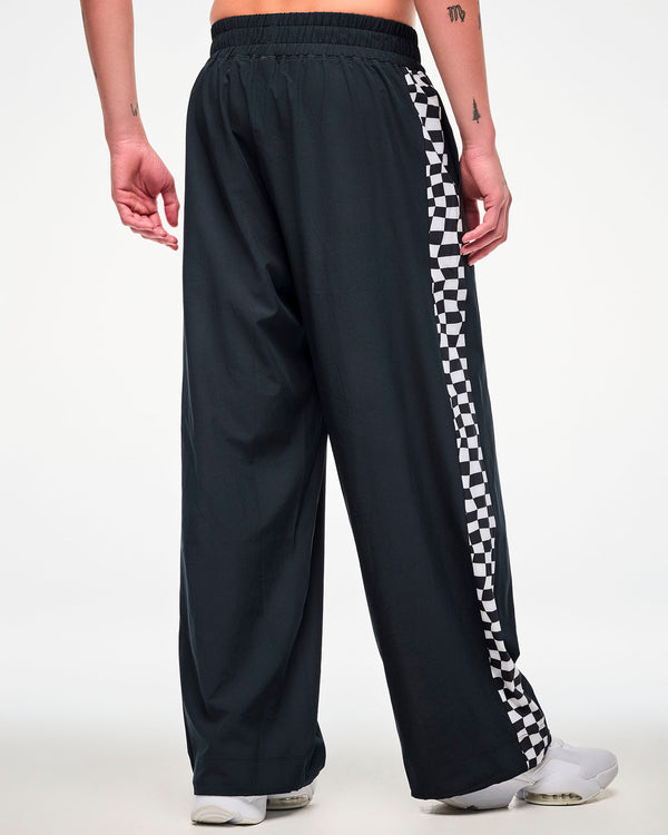 Back view of a male model wearing black track pants, checkerboard side panel and white sneakers.