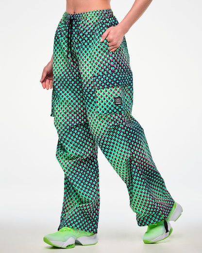 Female model with her hand in the side pocket of green cargo pants featuring an aqua, black, and pink dot print and a black tag with a cassette tape icon and 'ZUMBA' text on the left cargo pocket, paired with green sneakers.