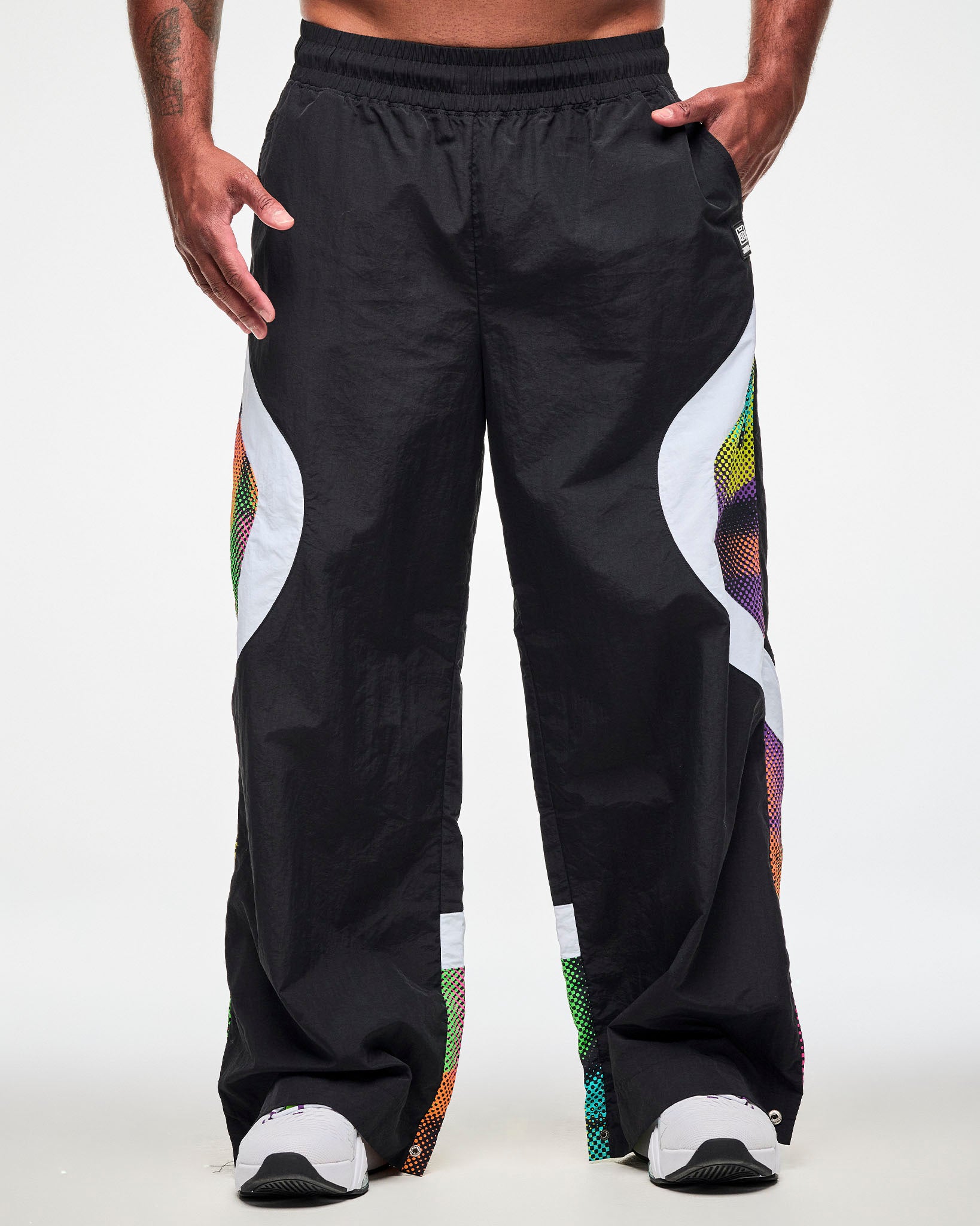 Male model wearing black track pants with a white and multicolor dot-print side panel and white sneakers, one hand in the side pockets.