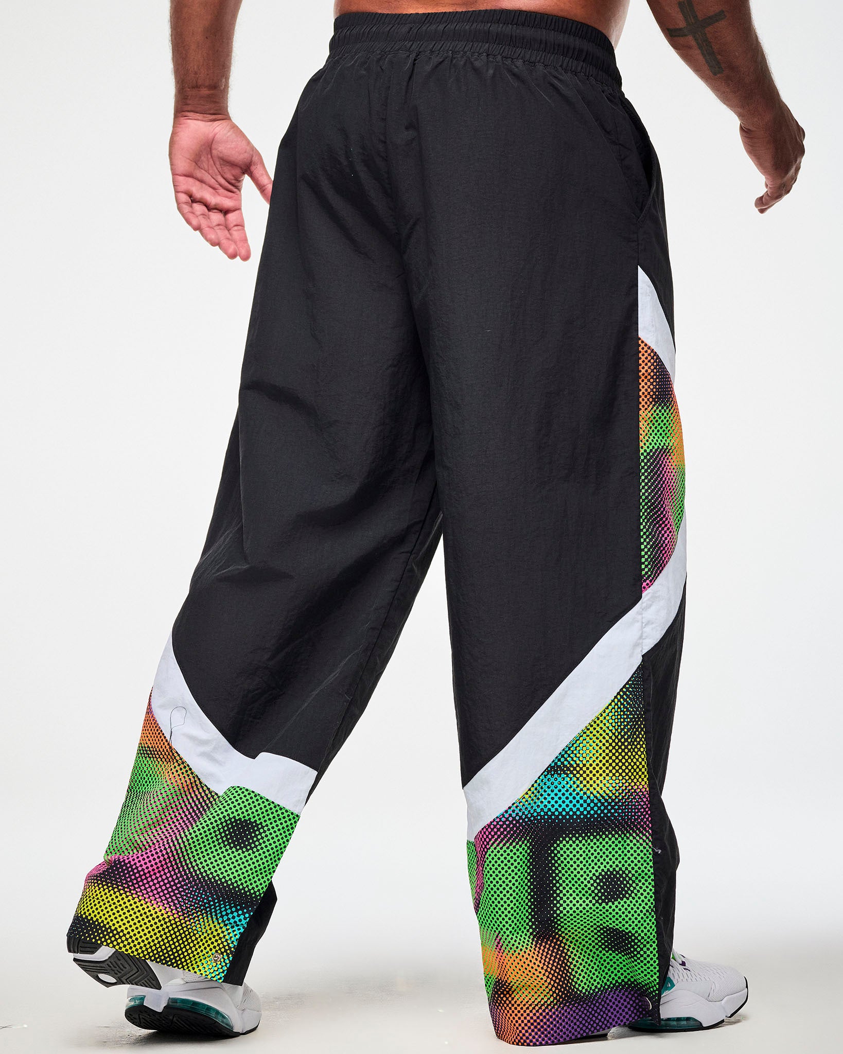 Shown from the back, male model wearing black track pants with a white and multicolor dot-print side panel and white sneakers.