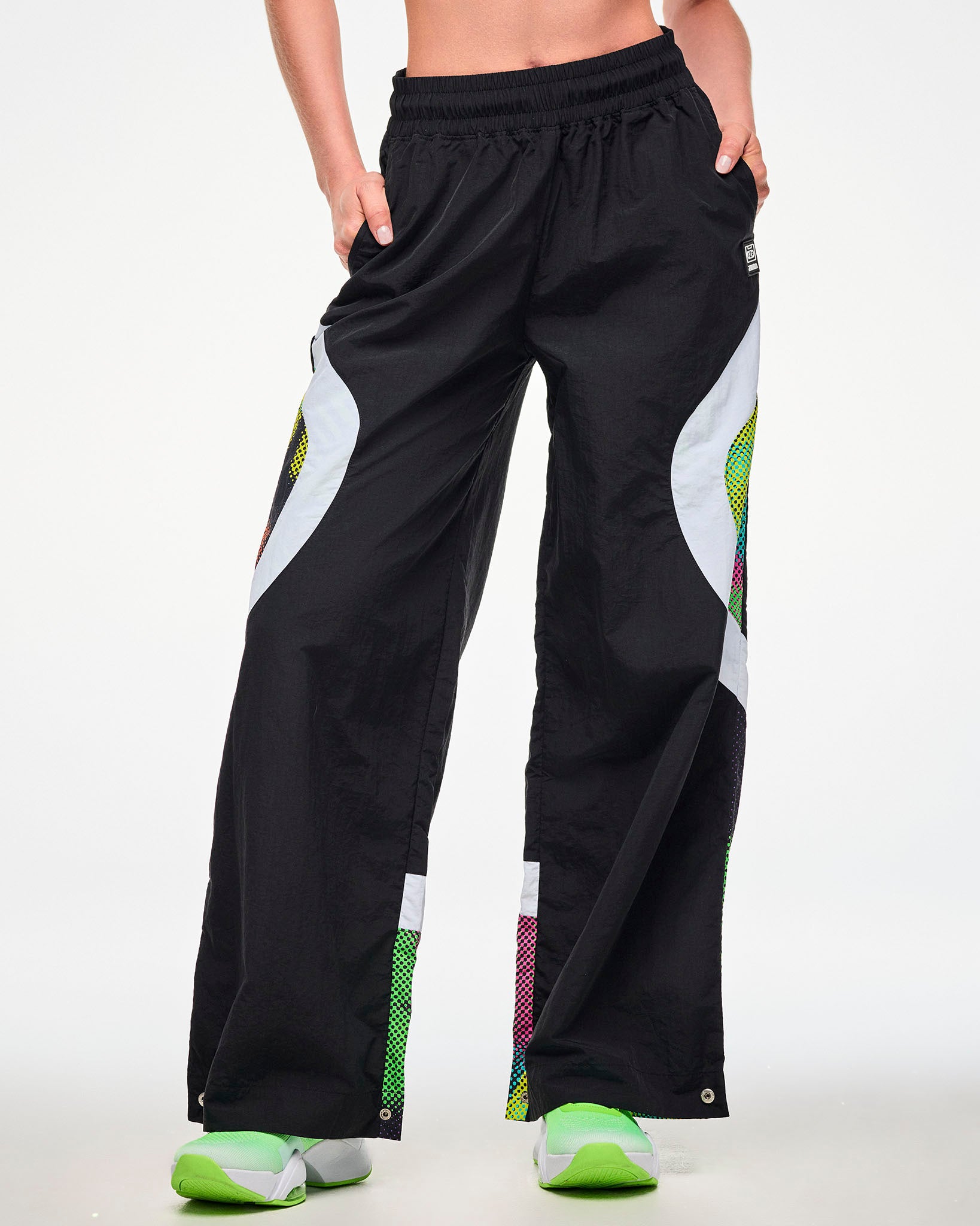 Female model wearing black track pants with a white and multicolor dot-print side panel and green sneakers, hand tucked into the side pocket.