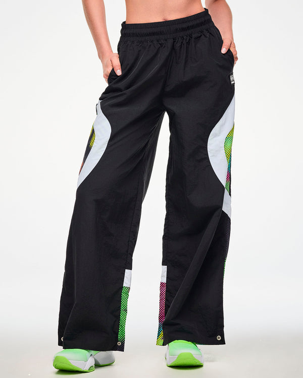 Female model wearing black track pants with a white and multicolor dot-print side panel and green sneakers, hand tucked into the side pocket.