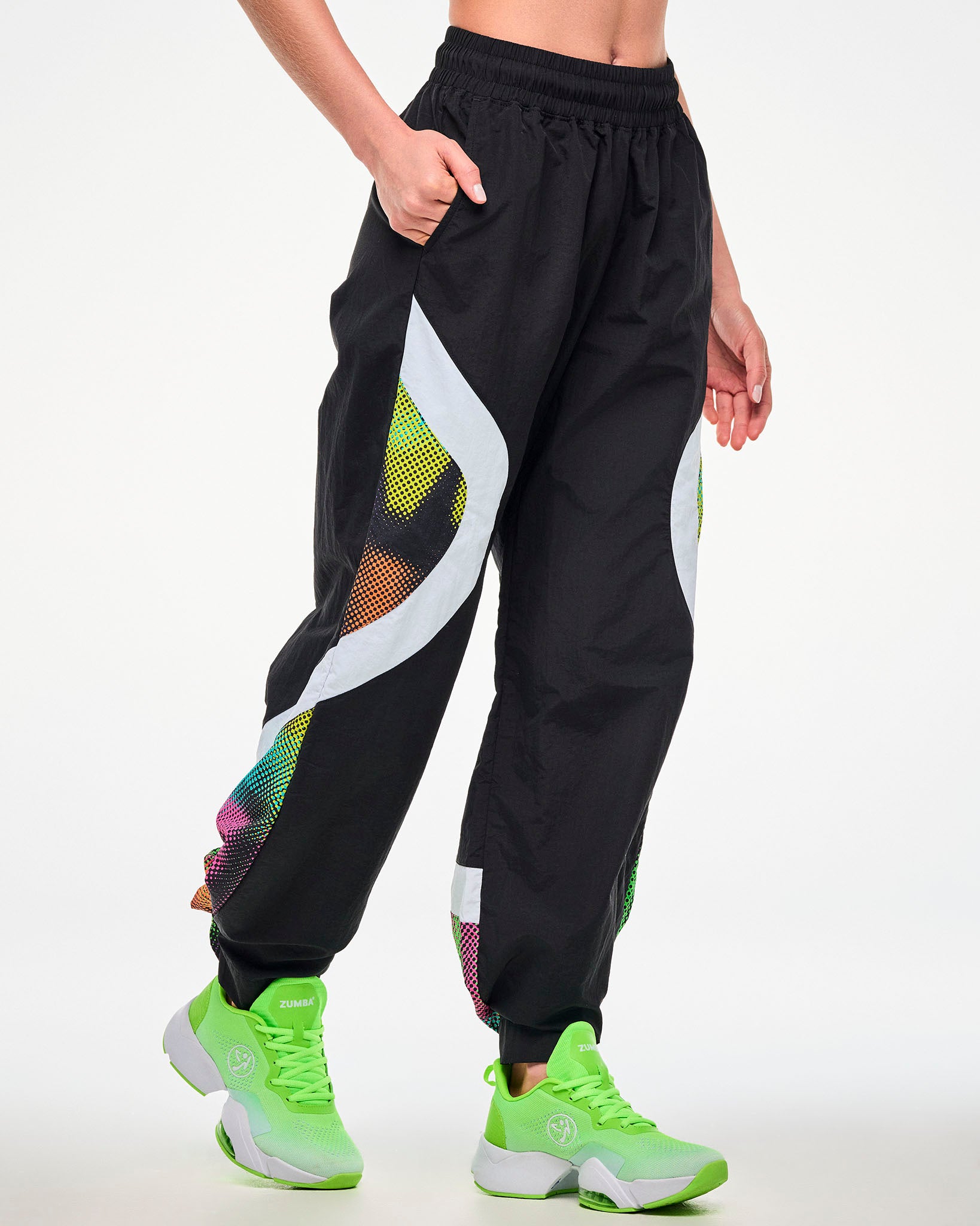 Female model wearing black track pants with a white and multicolor dot-print side panel, hand in the right pocket.