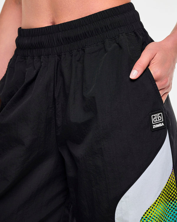 Close-up of a female model with a hand in the left pocket of black track pants, showing a black tag featuring a cassette tape icon and 