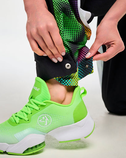 Close-up of a hand unsnapping the ankle closure on black track pants with a multicolor dot-print panel, paired with green sneakers.