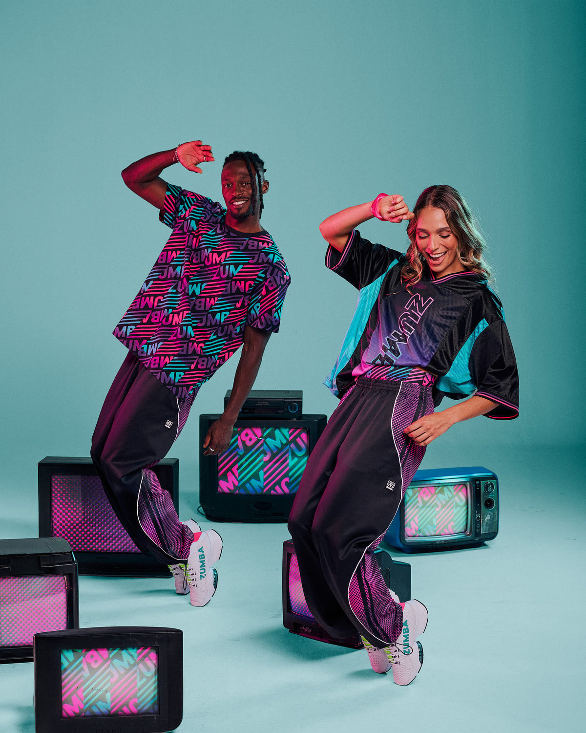 A male and female model dancing in matching streetwear outfits with retro TV props against a teal studio background. The outfits feature a black base with vibrant pink, purple, and aqua graphic patterns and "ZUMBA" text.