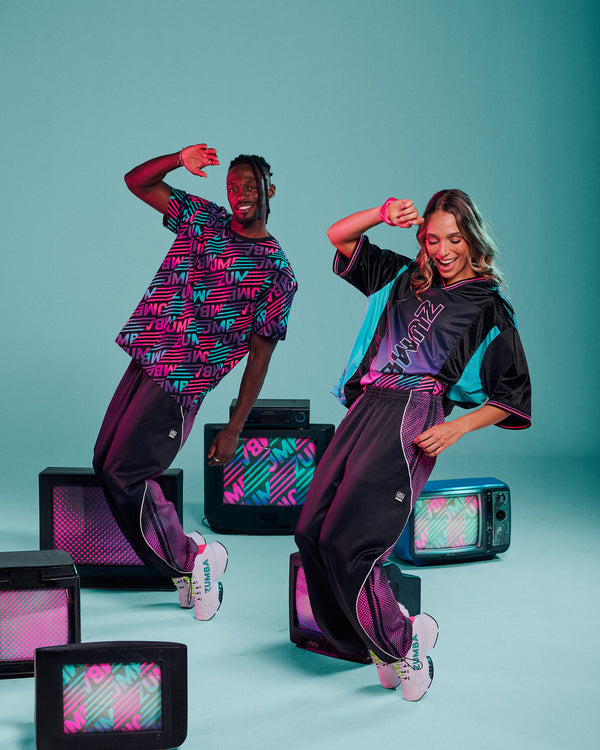 A male and female model dancing in matching streetwear outfits with retro TV props against a teal studio background. The outfits feature a black base with vibrant pink, purple, and aqua graphic patterns and 