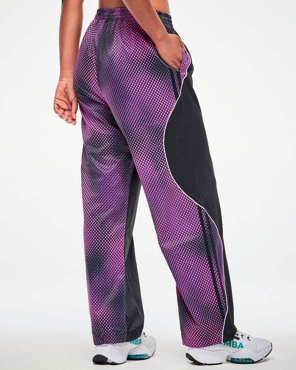 Back view of a female model wearing black track pants with a purple-to-pink dot gradient side and back panel, paired with white shoes.
