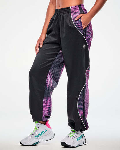 Female model wearing black track pants with a purple-to-pink dot gradient side and back panel, hand in the pocket, view from the side.