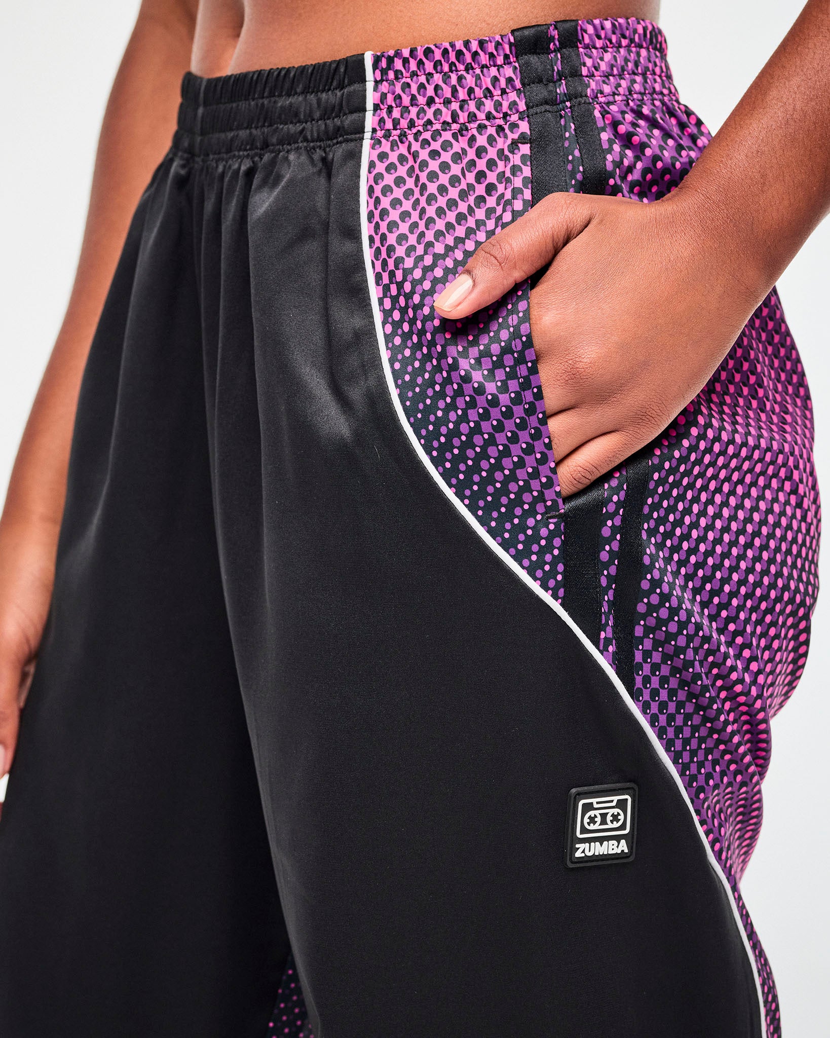 Close-up of the left side pocket on black track pants, where the purple-to-pink gradient panel meets the black front, with a black tag featuring a cassette tape icon and "ZUMBA" text, both in white, just below the pocket.