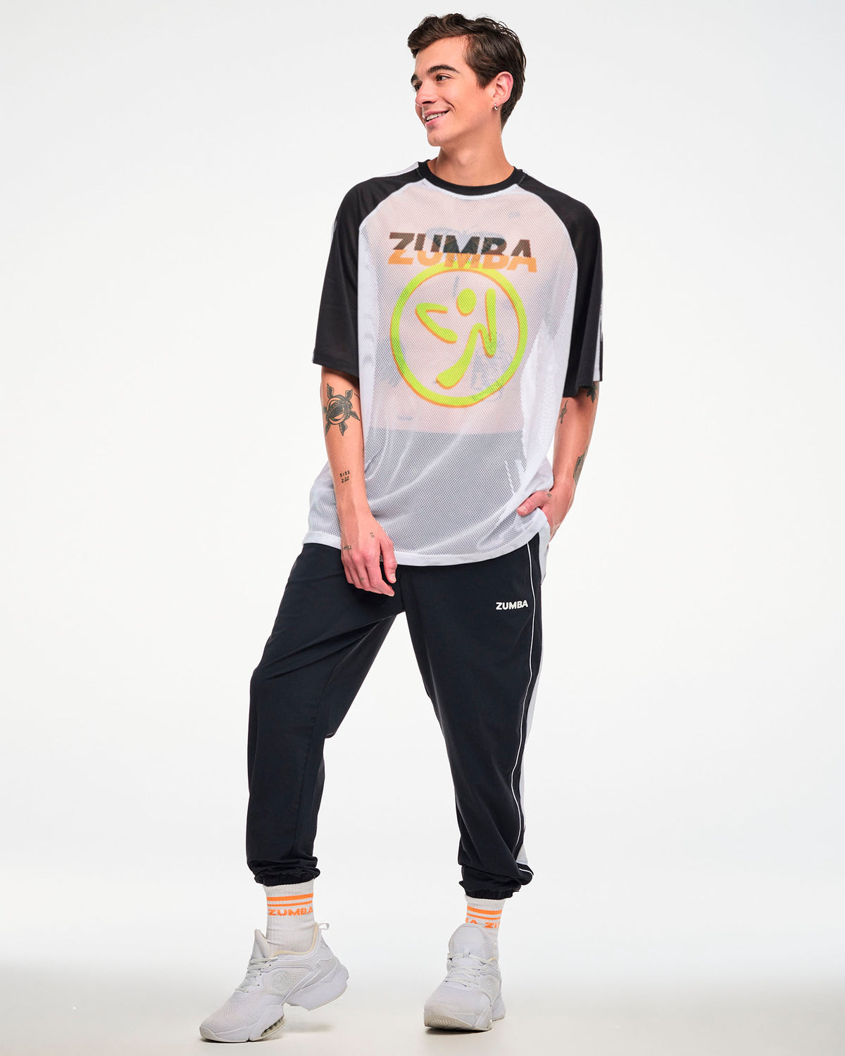 Male model, full view, wearing a white and black mesh tee with a yellow Zumbito logo, and "ZUMBA in black and orange text, paired with black joggers and white shoes.