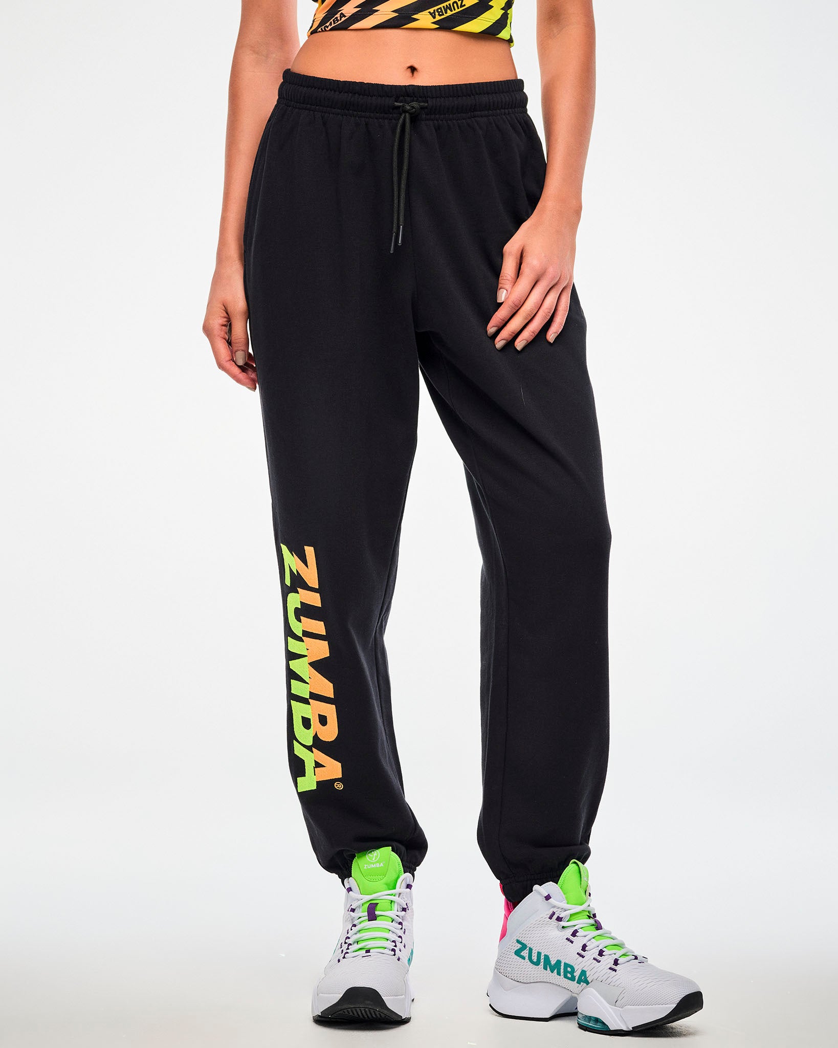 Close-up of a female model wearing black joggers with a green-to-orange gradient "ZUMBA" leg print and paired with white shoes.