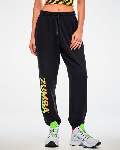 Close-up of a female model wearing black joggers with a green-to-orange gradient "ZUMBA" leg print and paired with white shoes.