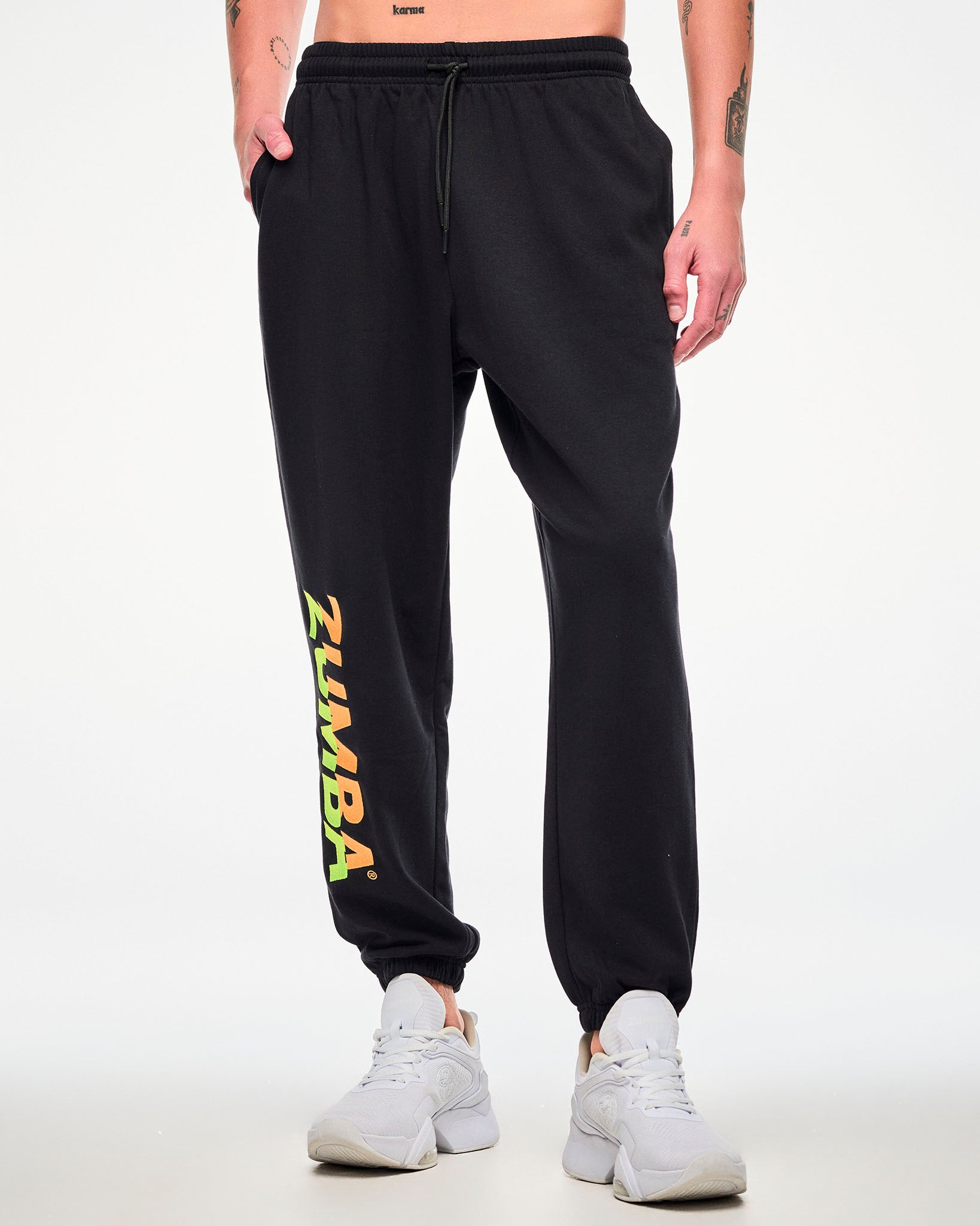 Male model wearing black joggers with a drawstring waist, hand in his pocket and a green-to-orange gradient "ZUMBA" leg print, paired with white sneakers.