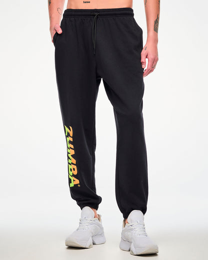 Male model wearing black joggers with a drawstring waist, hand in his pocket and a green-to-orange gradient "ZUMBA" leg print, paired with white sneakers.