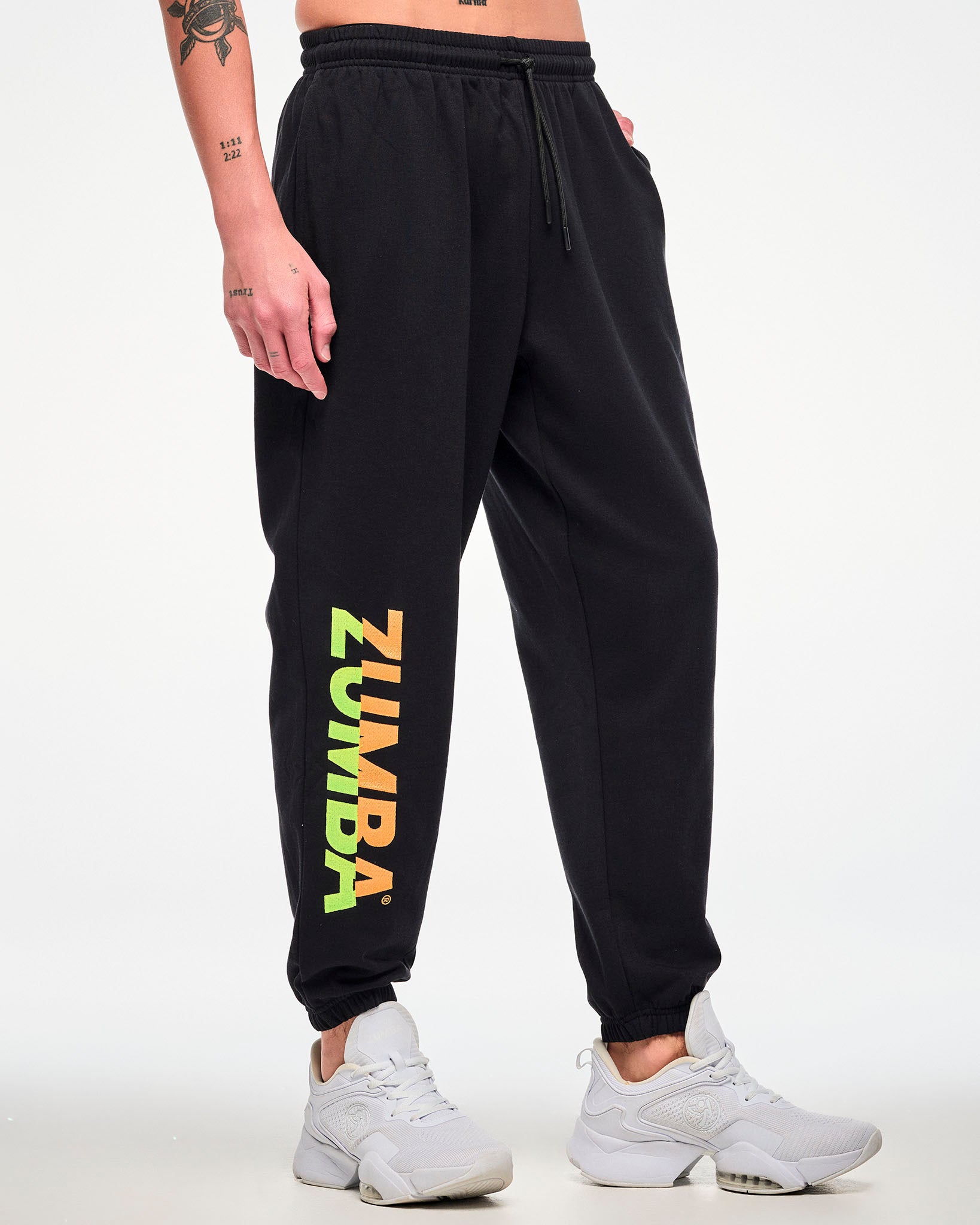Male model wearing black joggers with a green-to-orange gradient "ZUMBA" leg print and white sneakers.