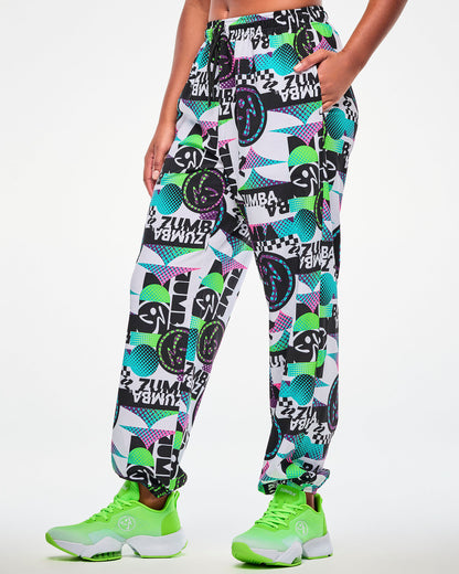 Female model wearing white track pants with an all-over abstract pattern of shapes and Zumbito logos in black, aqua, pink, and green, and black and white "ZUMBA" text, hand in the side pocket, paired with green sneakers.