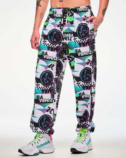 Male model wearing white track pants with an all-over abstract pattern of shapes and Zumbito logos in black, aqua, pink, and green, and black and white "ZUMBA" text, hand in the pocket, paired with white sneakers.