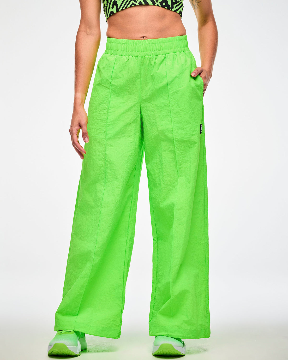 Female model wearing green wide-leg track pants and green shoes, hand in the side pocket.