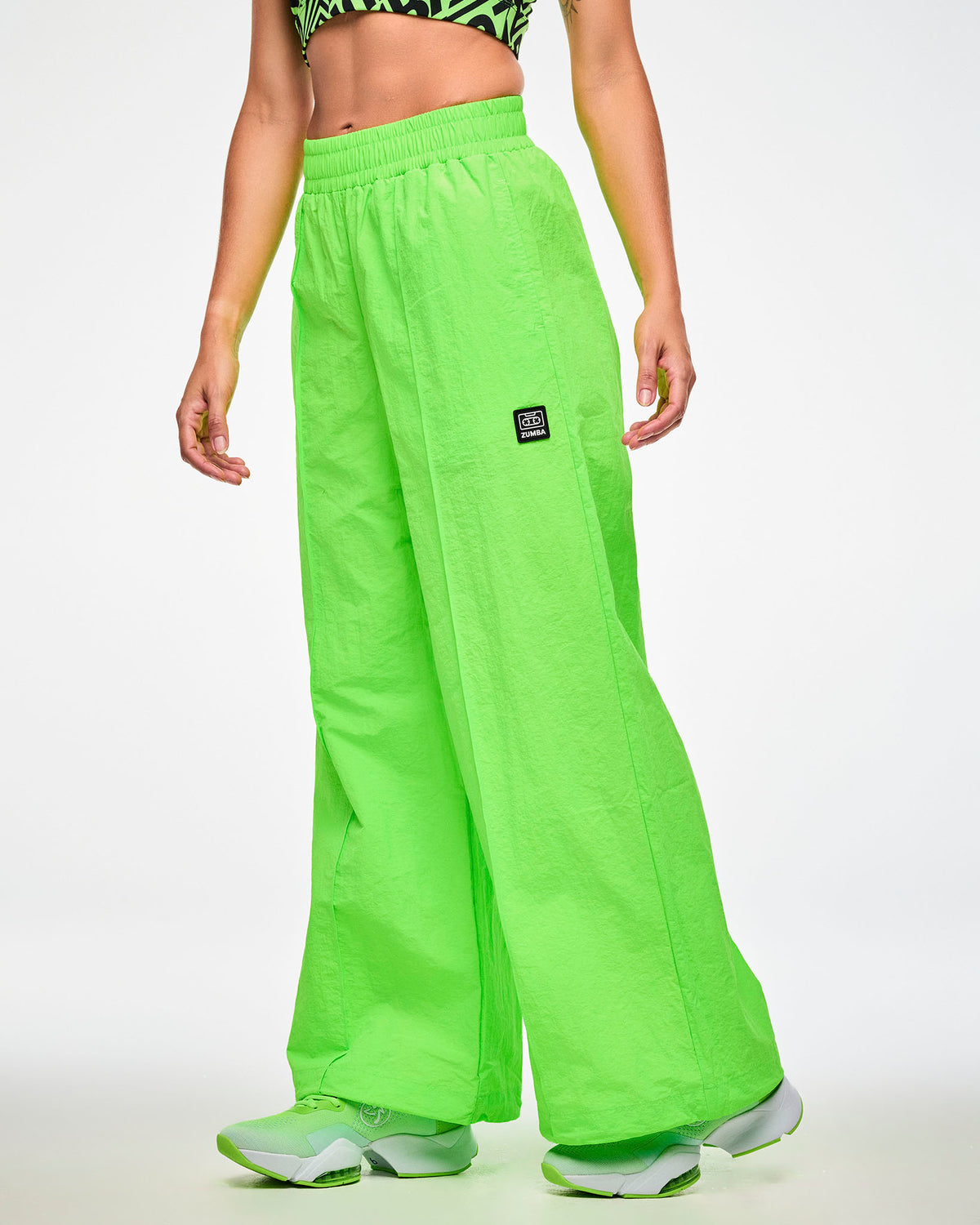 Female model wearing green wide-leg track pants with a cassette tag patch on the upper left leg and paired with matching green shoes.