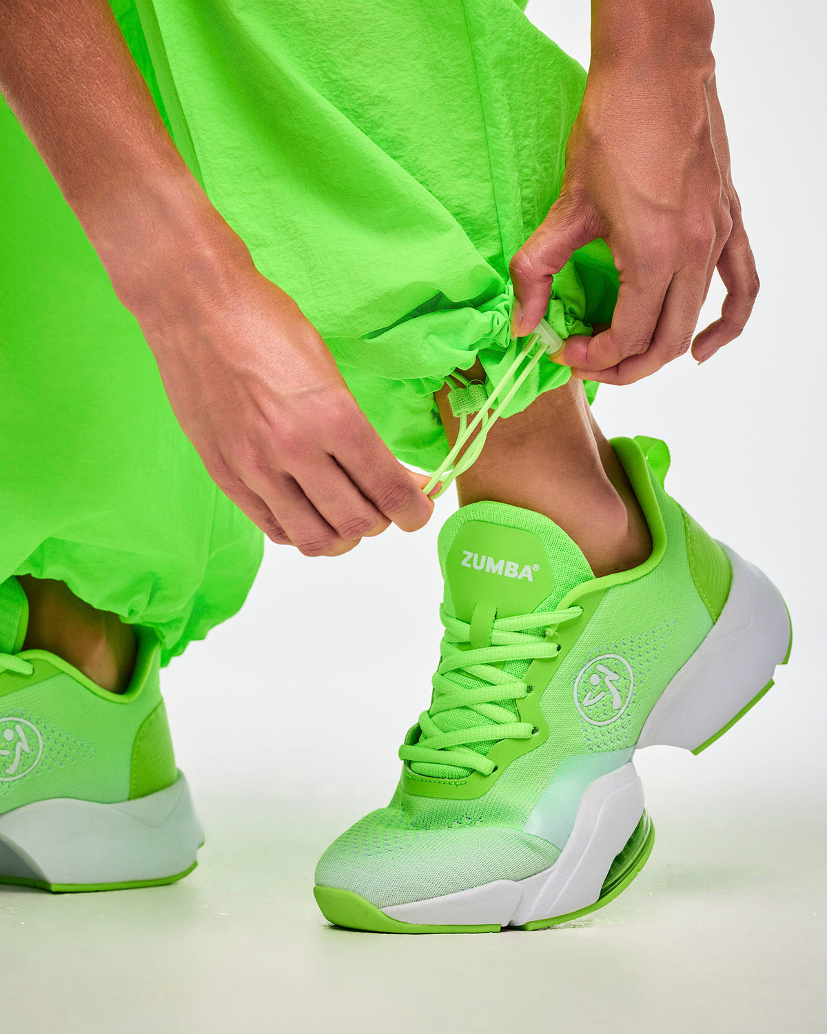 Close-up of hands cinching the bungee cord along the ankle cuff on green track pants and paired with green shoes.

