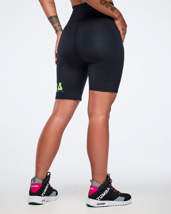 Back view of a female model wearing black biker shorts with a logo and Zumba shoes on a white background.