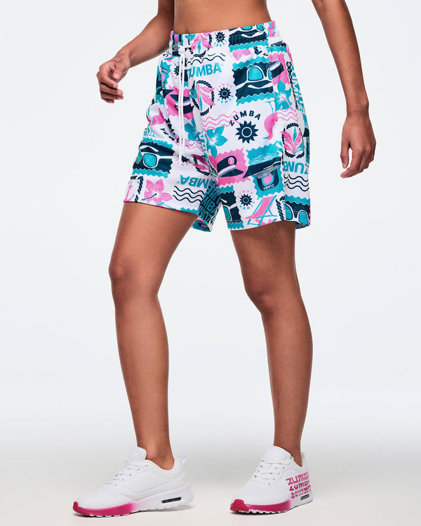 Side view of white elastic-waist drawstring shorts with a teal-and-pink Zumba seaside collage print and front pockets; shown on a female model with white sneakers.