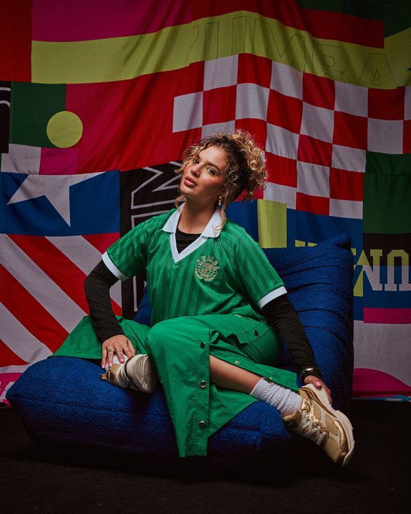 Female model wearing a green polo shirt and green side snap sitting on a blue bean bag chair against a colorful geometric-patterned background.