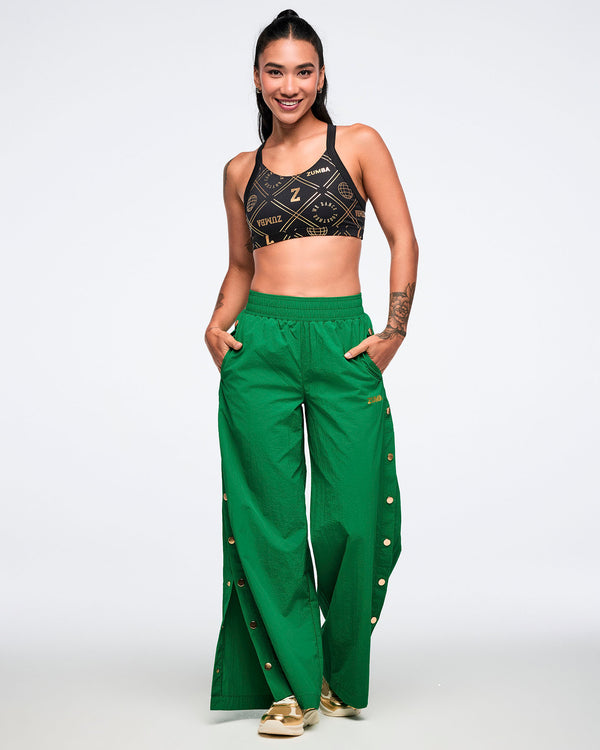 Woman wearing a black sports bra with gold accents and green high-waisted side snap pants on a white background.