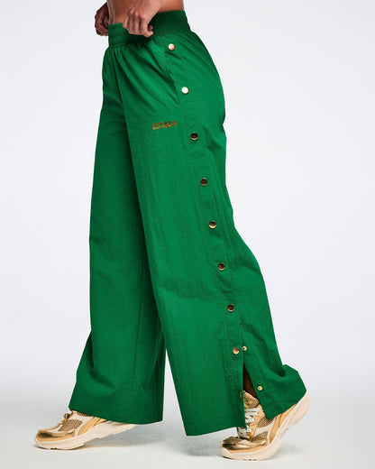Female model wearing green wide-leg pants with gold side snaps and gold shoes, on a white background.