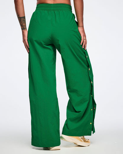 Female model wearing green wide-leg pants, shown from the back, with gold side snaps and gold shoes, on a white background.