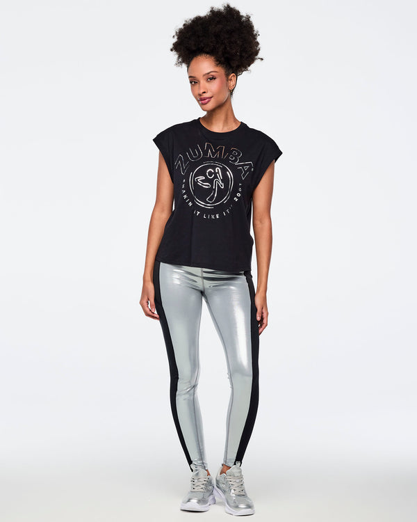 Metallic silver high-waisted leggings with black side panels. Female model wears a black sleeveless Zumba graphic tee, shiny silver full-length leggings with a wide waistband and contrast side panels, and silver athletic sneakers, standing front-facing against a light gray background.