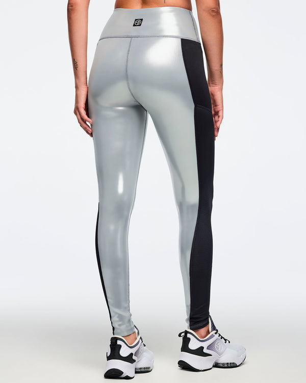 Metallic silver high-waisted leggings with black side panels and a small back waistband logo. Female model wears fitted glossy full-length leggings with a wide waistband and white athletic sneakers, shown from the back against a light gray background.
