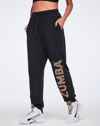 Side view of black sweatpants with an elastic drawstring waistband, tapered leg, side pockets, and leopard-print “ZUMBA” graphic down one leg. Female model wears the pants with white high-top Zumba sneakers.