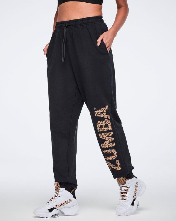 Side view of black sweatpants with an elastic drawstring waistband, tapered leg, side pockets, and leopard-print “ZUMBA” graphic down one leg. Female model wears the pants with white high-top Zumba sneakers.