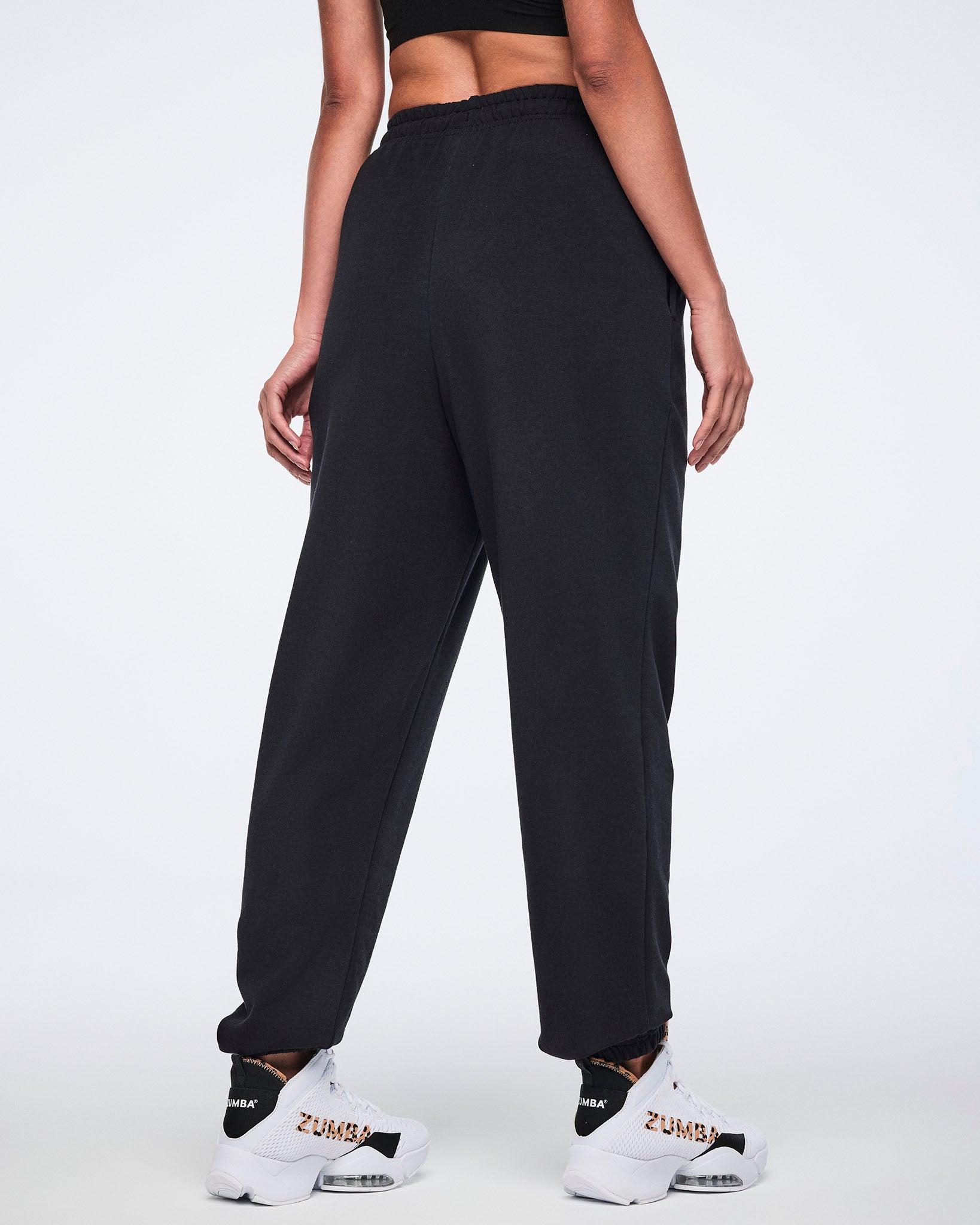 Back view of black sweatpants with an elastic waistband, tapered leg, and gathered cuffs. Female model wears the pants with white high-top Zumba sneakers.