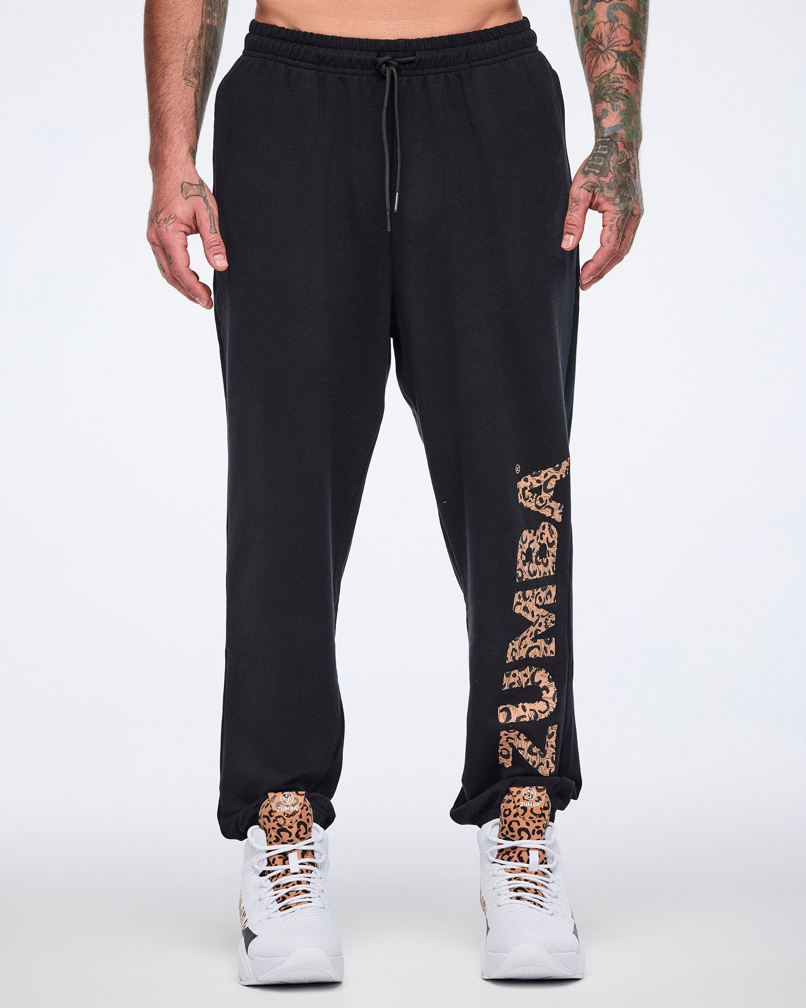 Front view of black sweatpants with an elastic drawstring waistband, tapered leg, and leopard-print “ZUMBA” graphic down the left leg. Male model wears the pants with white high-top Zumba sneakers.