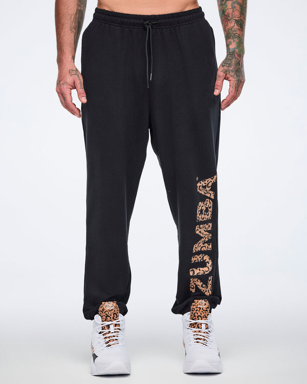 Front view of black sweatpants with an elastic drawstring waistband, tapered leg, and leopard-print “ZUMBA” graphic down the left leg. Male model wears the pants with white high-top Zumba sneakers.