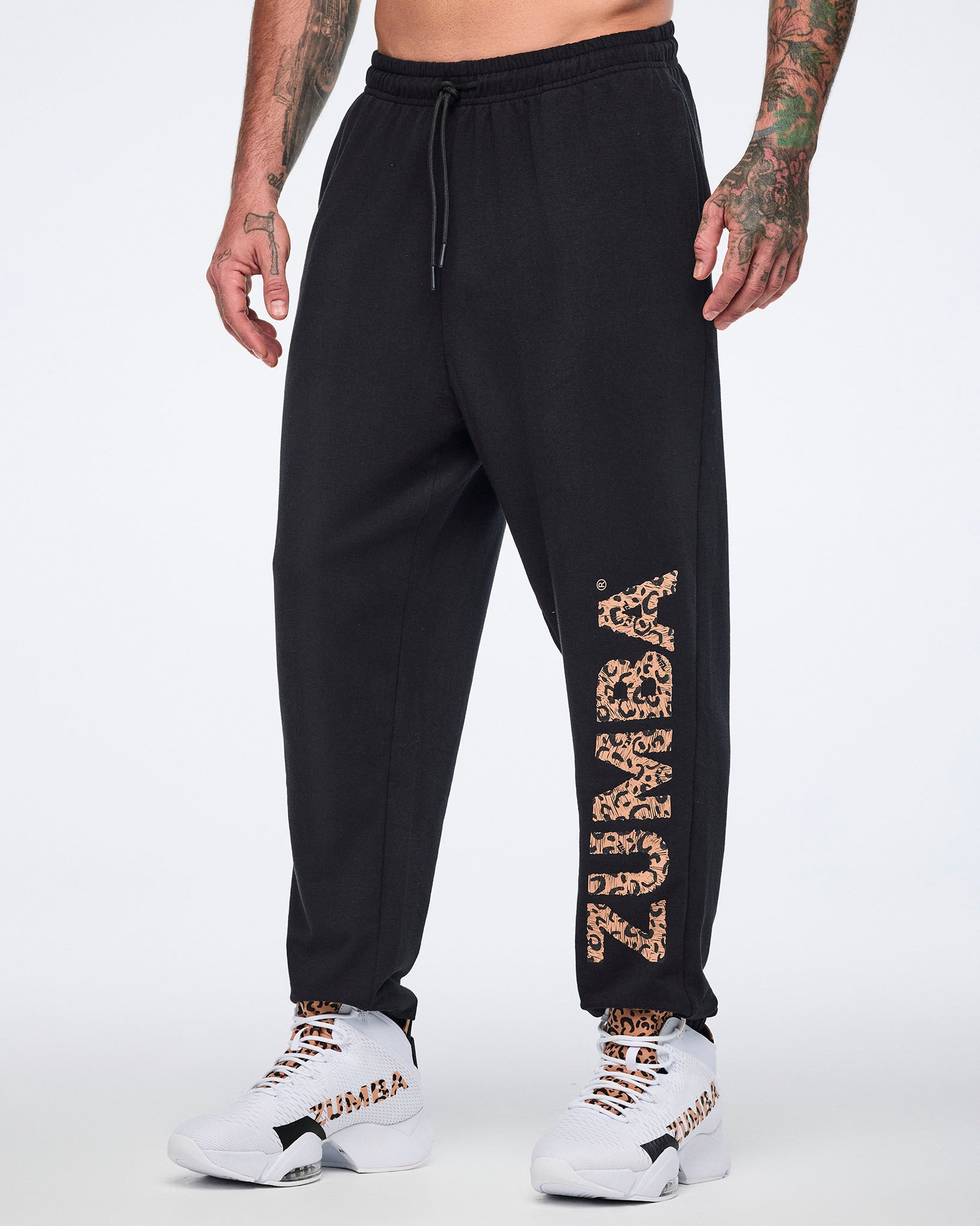 Angled front view of black sweatpants with an elastic drawstring waistband, tapered leg, and leopard-print “ZUMBA” graphic down the left leg. Male model wears the pants with white high-top Zumba sneakers.