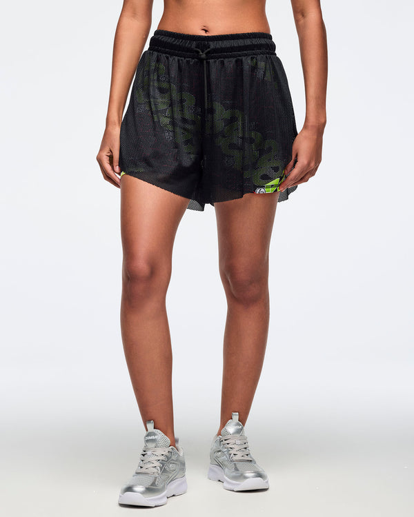 Female model wears black mesh overlay shorts with a colorful printed inner short and drawstring waistband, layered over fitted multicolor inner shorts, paired with silver athletic sneakers, shown in a front view against a light gray background.