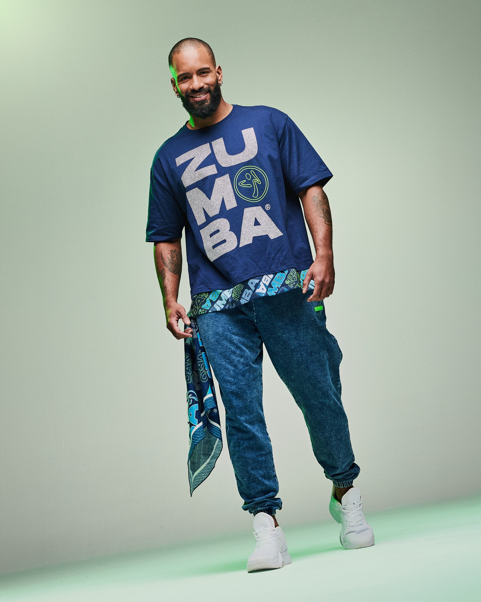 Male model wearing a blue “Zumba” shirt with blue denim pants and a patterned bandana hanging from his pocket, paired with white shoes against a light green background.