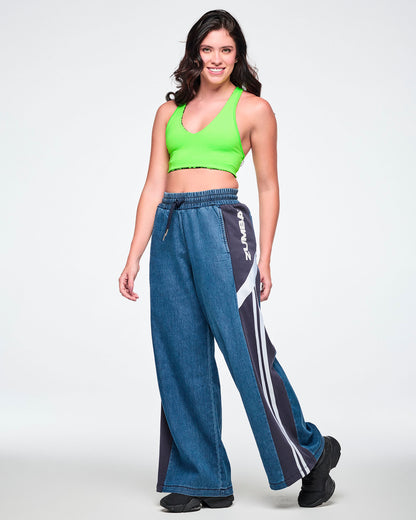 Female model wearing a neon green bra top with wide-leg combo pants featuring a navy side panel, white "ZUMBA" text, a diagonal white stripe, and two vertical white stripes down the leg, paired with black sneakers.