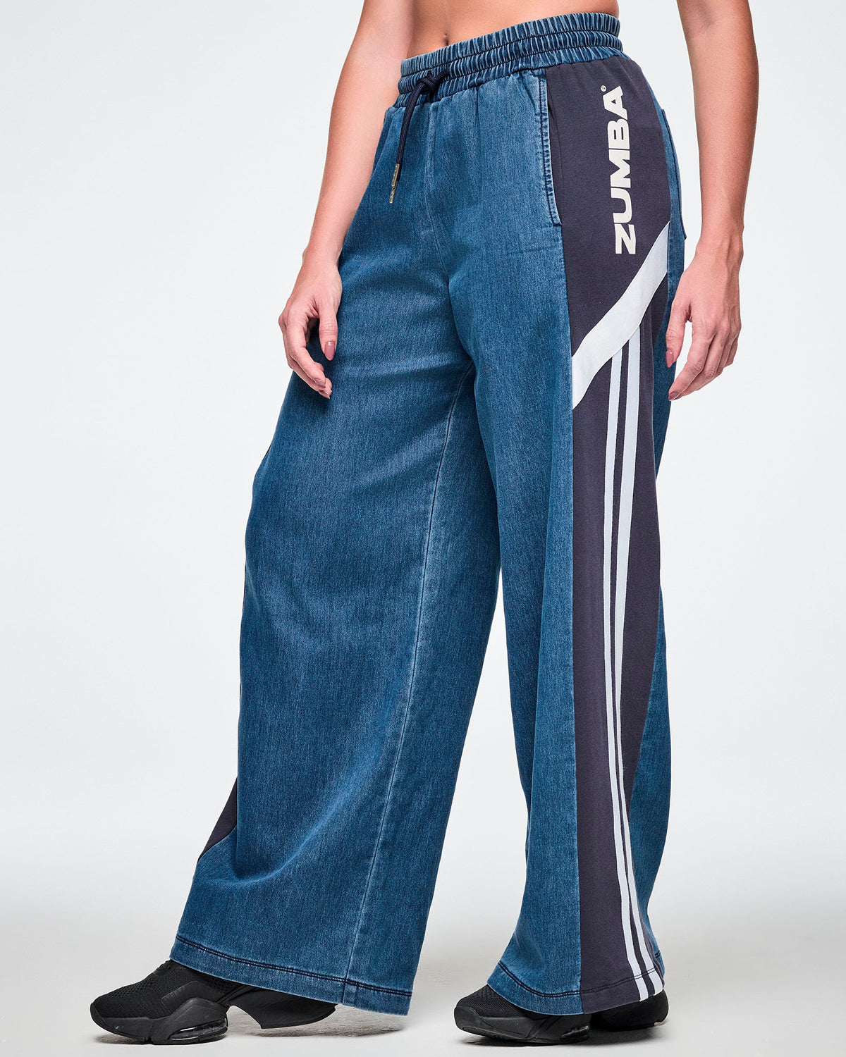 Side view close-up of a female model wearing wide-leg combo pants, showing the elastic drawstring waistband and the navy side panel with white "ZUMBA" text, a diagonal stripe, and two vertical stripes running down the leg.