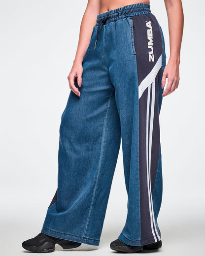 Side view close-up of a female model wearing wide-leg combo pants, showing the elastic drawstring waistband and the navy side panel with white "ZUMBA" text, a diagonal stripe, and two vertical stripes running down the leg.