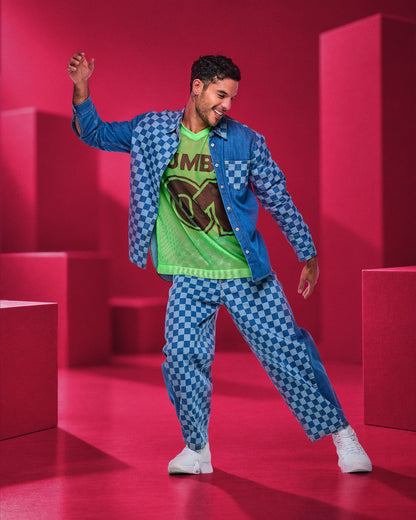Male model dancing in blue checkerboard denim barrel ankle pants, an open checkerboard denim shirt, and a neon green oversized mesh V-neck top, surrounded by pink blocks.
