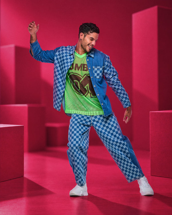 Male model dancing in blue checkerboard denim barrel ankle pants, an open checkerboard denim shirt, and a neon green oversized mesh V-neck top, surrounded by pink blocks.